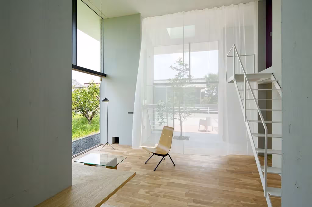 Espacio de doble altura en la casa Ohno con cortina blanca, mesa de vidrio y vistas a los caquis