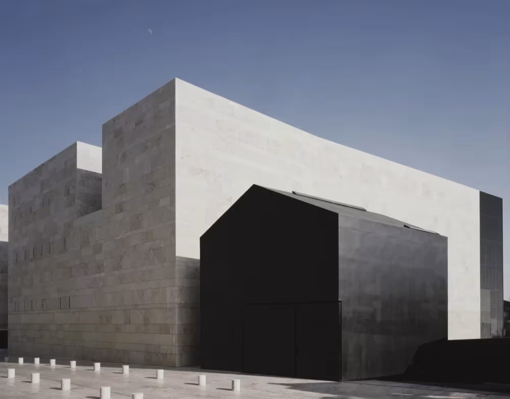 Esquina del Centro de Artes de Sines con volúmenes de piedra gris y una casa de silueta oscura en primer plano