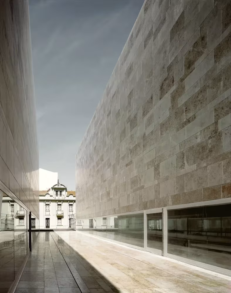 Calle entre el muro de piedra del Centro de Artes de Sines y una fachada de vidrio, con un edificio histórico al fondo