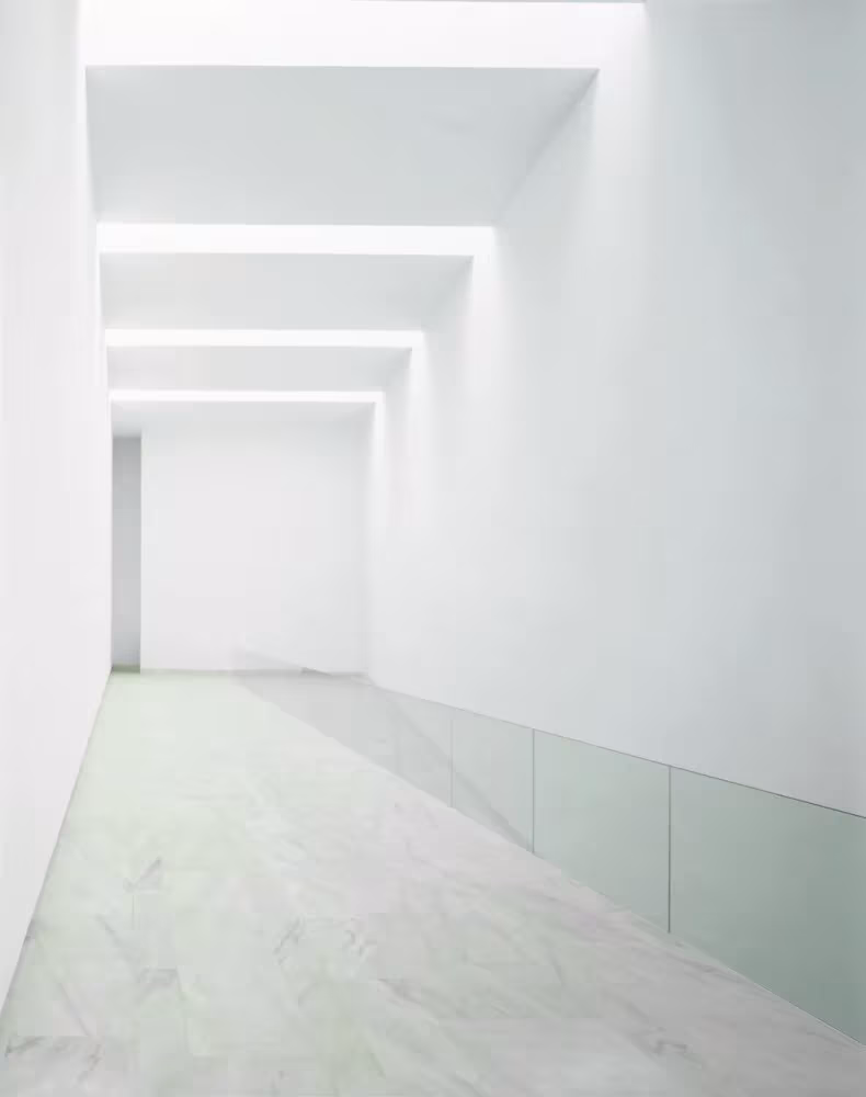 Pasillo blanco con rampa y barandilla de vidrio iluminado por una luz cenital lineal en el Centro de Artes de Sines