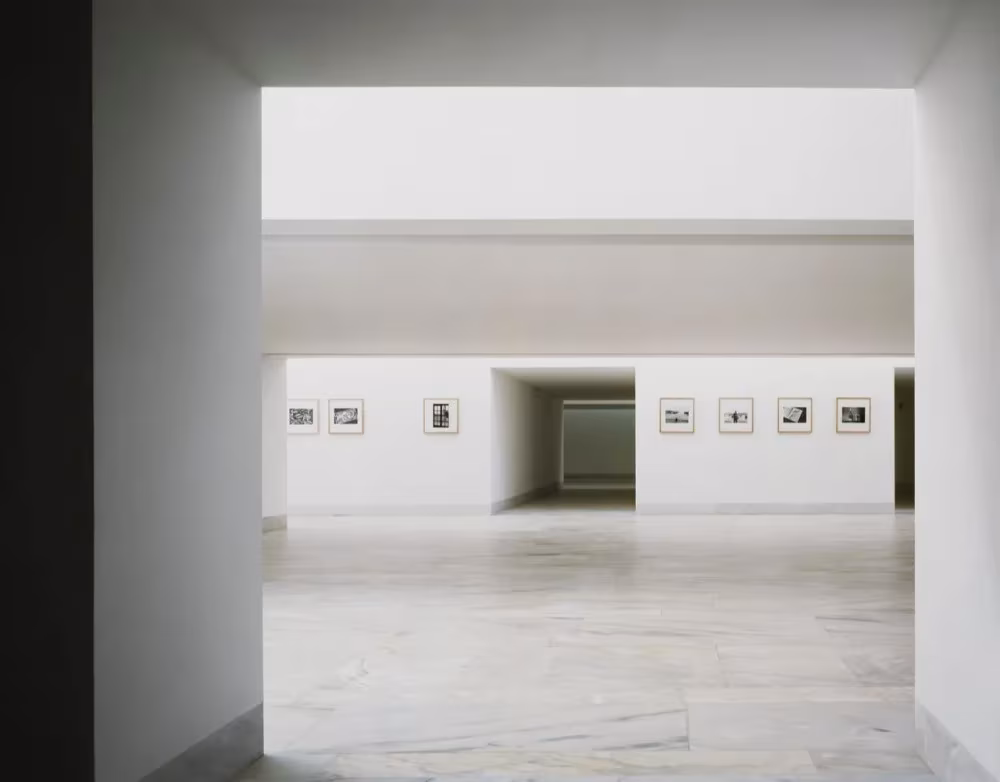 Vano blanco que da paso a una sala de exposiciones con fotografías colgadas y suelo de mármol en Sines