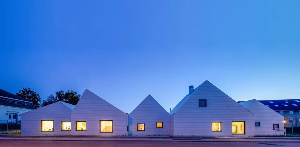 Fachada alargada de Livsrum al anochecer, con sus tejados a dos aguas blancos y ventanas iluminadas