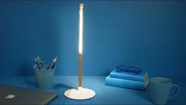 Lámpara de pie fina encendida sobre un escritorio azul con libros, taza y gafas
