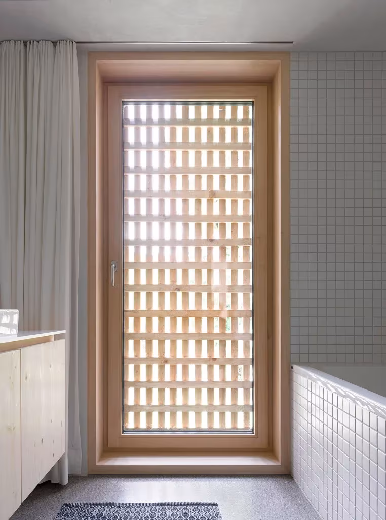Baño de azulejo blanco con puerta de celosía de madera y cortina gris en la Casa de Julia y Björn