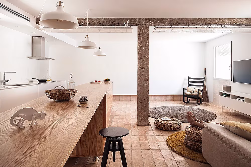 Salón cocina de L'Àtic Vernacle con isla de madera, pilar de hormigón, suelo de barro y cojines de esparto