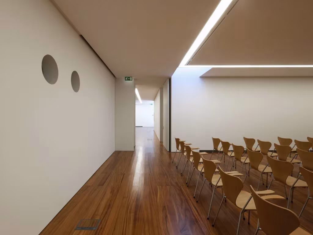 Auditorio del Espacio Miguel Torga con sillas de madera, óculos circulares en la pared y luz cenital