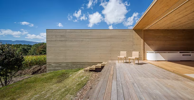 Terraza de madera con dos sillas bajo el voladizo de la Solid Cedar House frente al bosque y las nubes