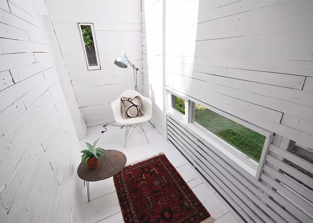 Estudio de madera pintada de blanco con alfombra roja, butaca y ventana horizontal al jardín