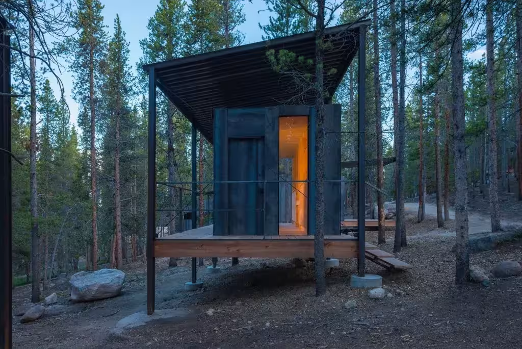 Cabaña de madera oscura sobre plataforma elevada bajo cubierta metálica en un bosque de Colorado