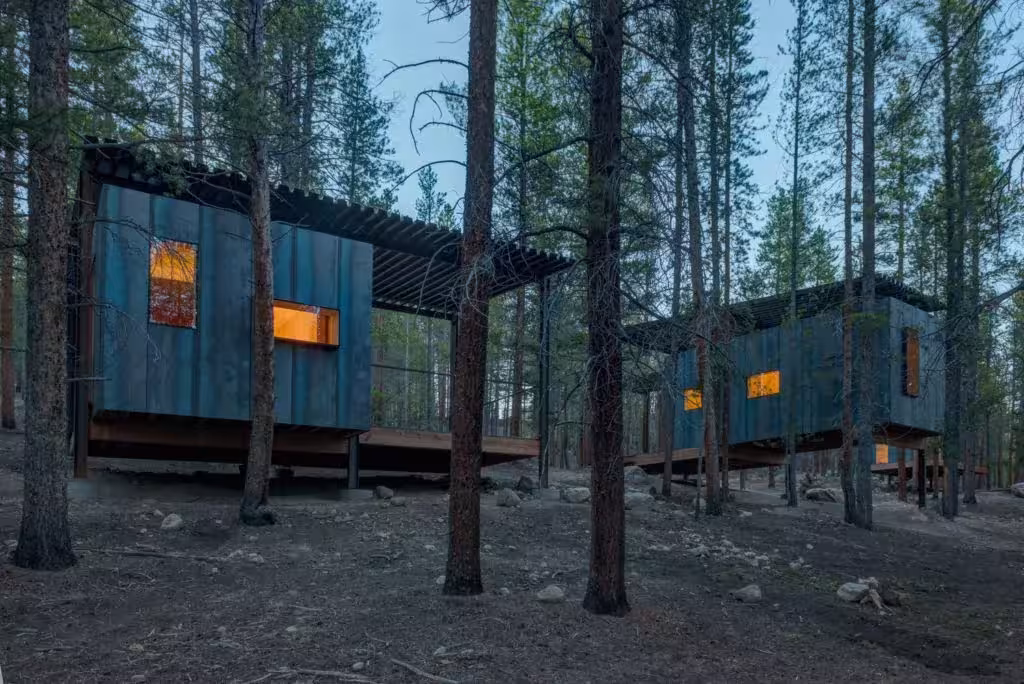 Dos cabañas diminutas de madera oscura con ventanas iluminadas en el bosque de Colorado Outward Bound