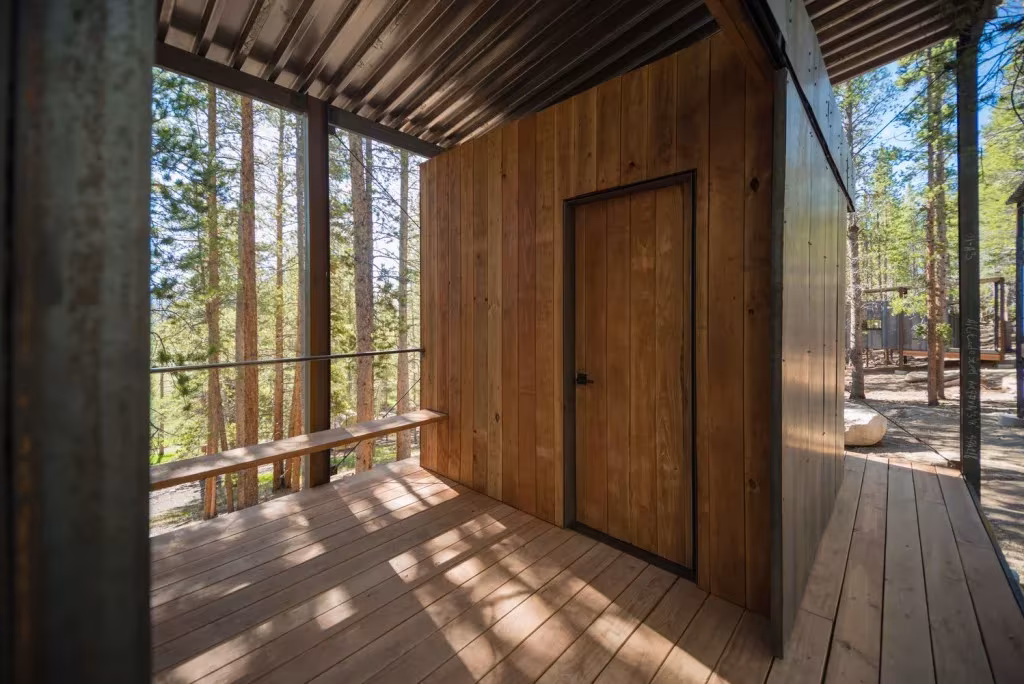 Porche de tablas de madera bajo cubierta junto a la puerta de una cabaña, con luz filtrada entre los pinos