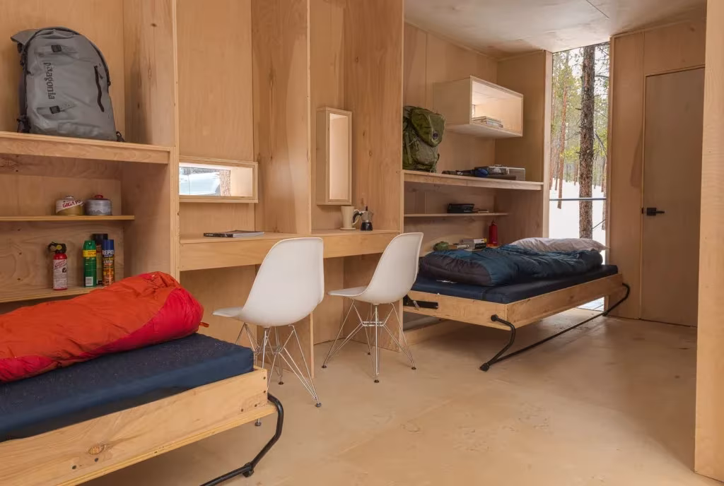 Interior de contrachapado de una cabaña con dos camas abatibles, escritorios y sillas blancas
