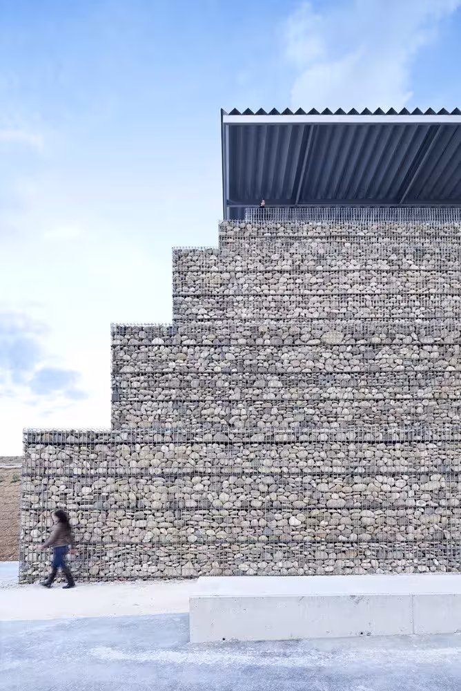 Muro escalonado de gaviones del Parque Venecia coronado por una pérgola metálica y una persona paseando