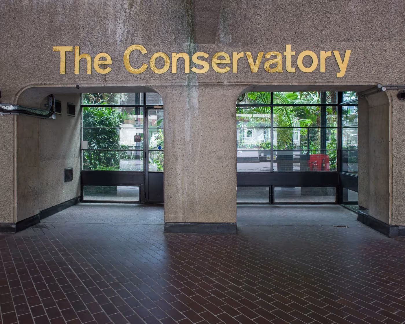 Entrada al Barbican Conservatory con el rótulo dorado, el pavimento de ladrillo y el hormigón brutalista, Londres