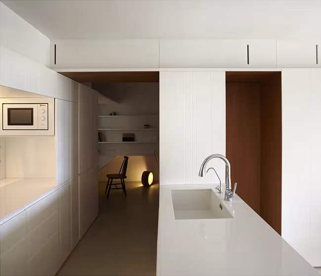 Cocina blanca de la Casa BHMM con grifo sobre la encimera y estancia con silla al fondo