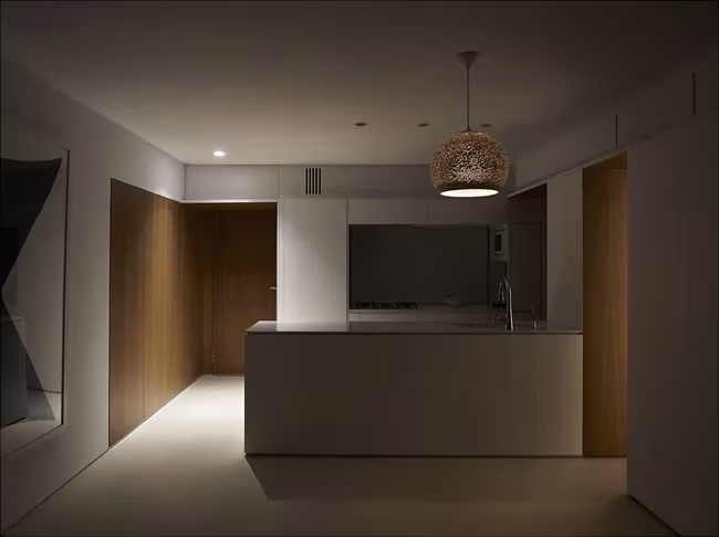 Cocina en penumbra de la Casa BHMM con isla blanca, lámpara colgante de fibra y paneles de madera