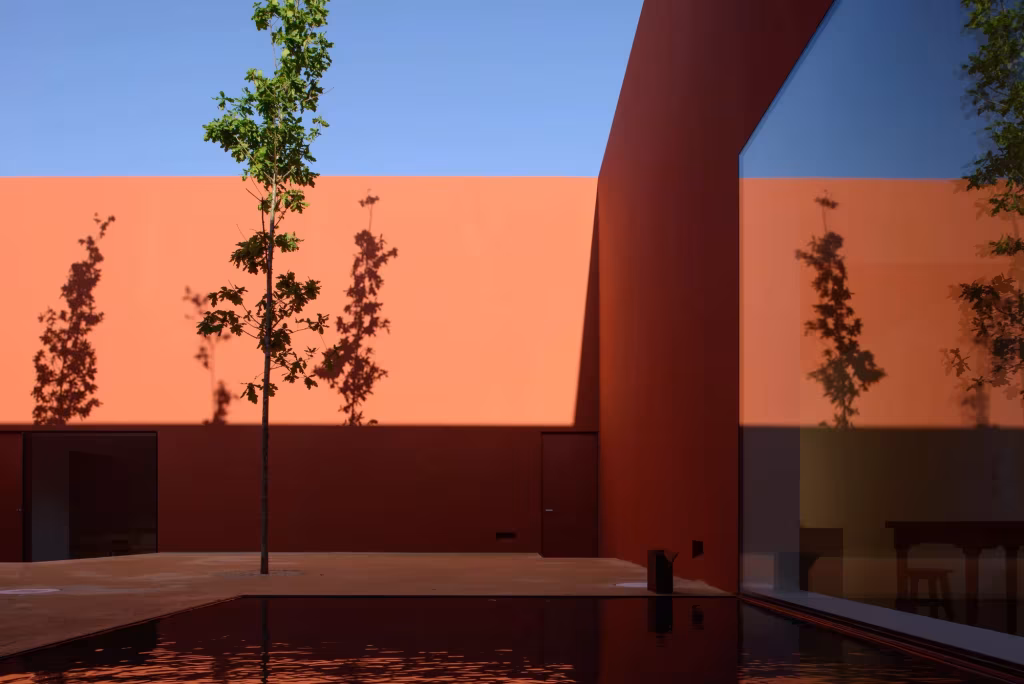 Patio de la Casa en Oeiras con muros naranja y rojo, árboles finos y lámina de agua al atardecer