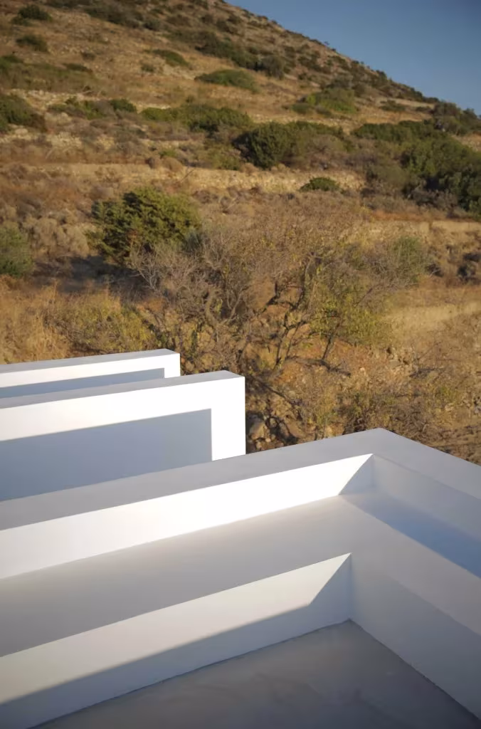 Muros blancos escalonados de la Casa Kamari frente a una ladera de matorral seco en Paros