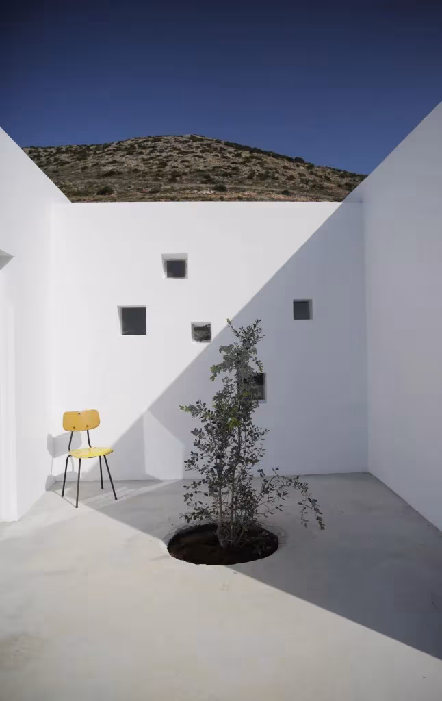 Patio blanco de la Casa Kamari con un olivo joven, una silla amarilla y pequeños huecos en el muro