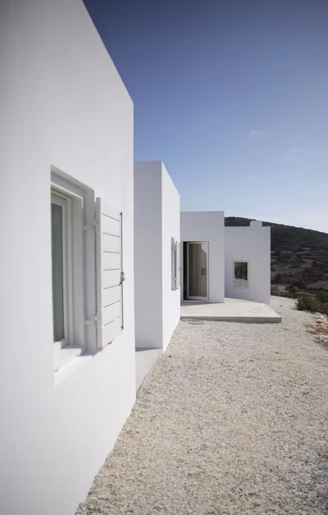 Fachada encalada de la Casa Kamari con contraventanas de madera y un camino de grava junto al muro