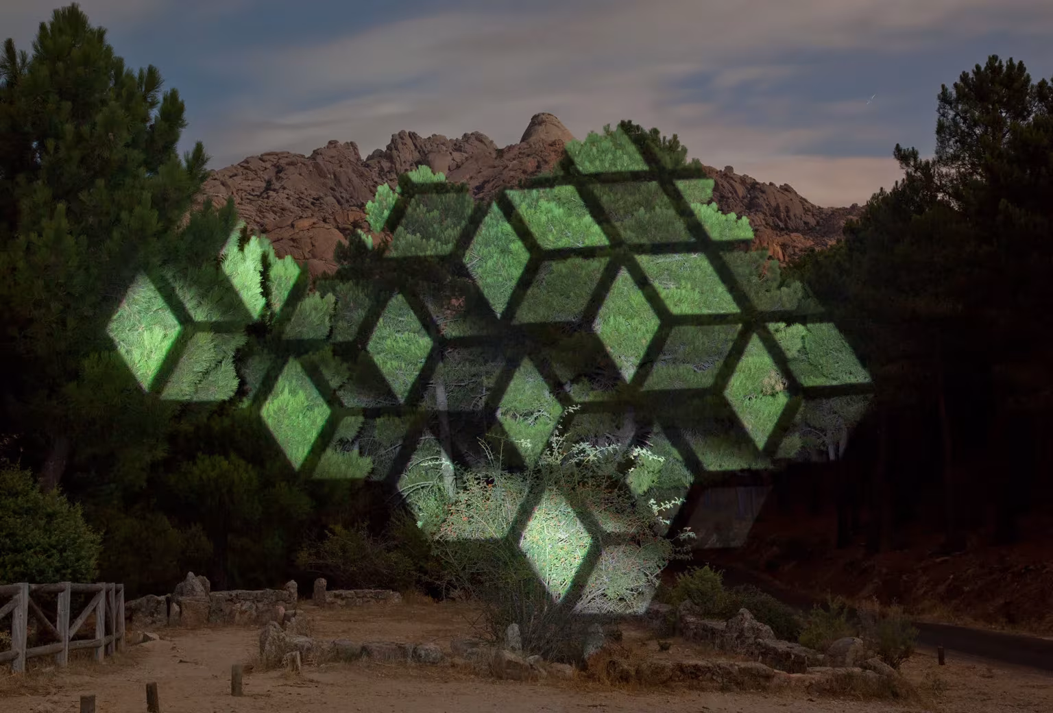 Intervención de luz de Javier Riera que proyecta hexágonos verdes sobre árboles en un paisaje rocoso al anochecer