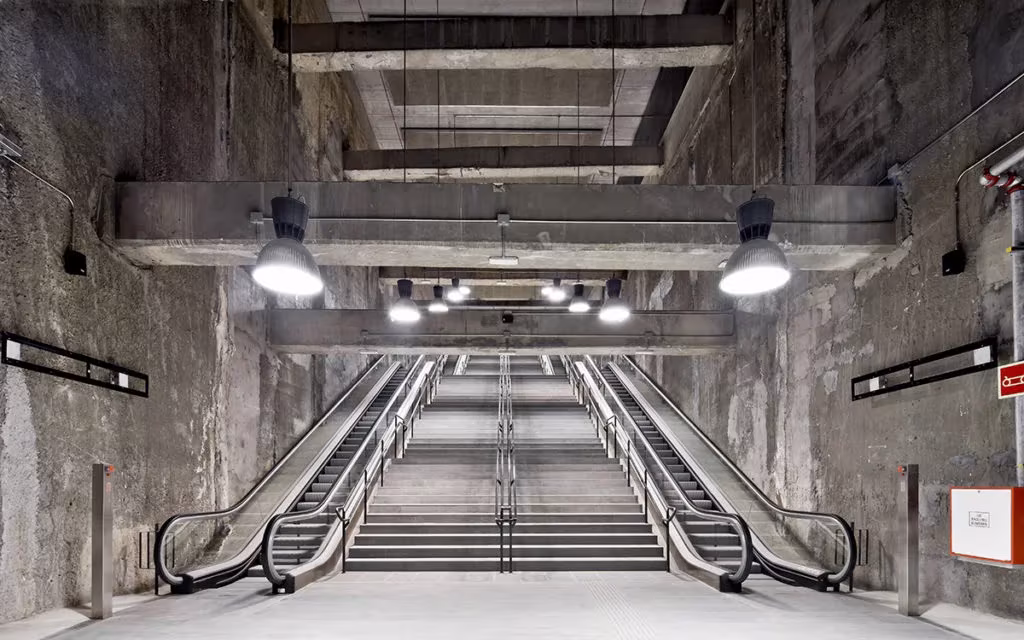 Escalera fija entre dos escaleras mecánicas bajo lámparas industriales en una estación de la L9 de Barcelona
