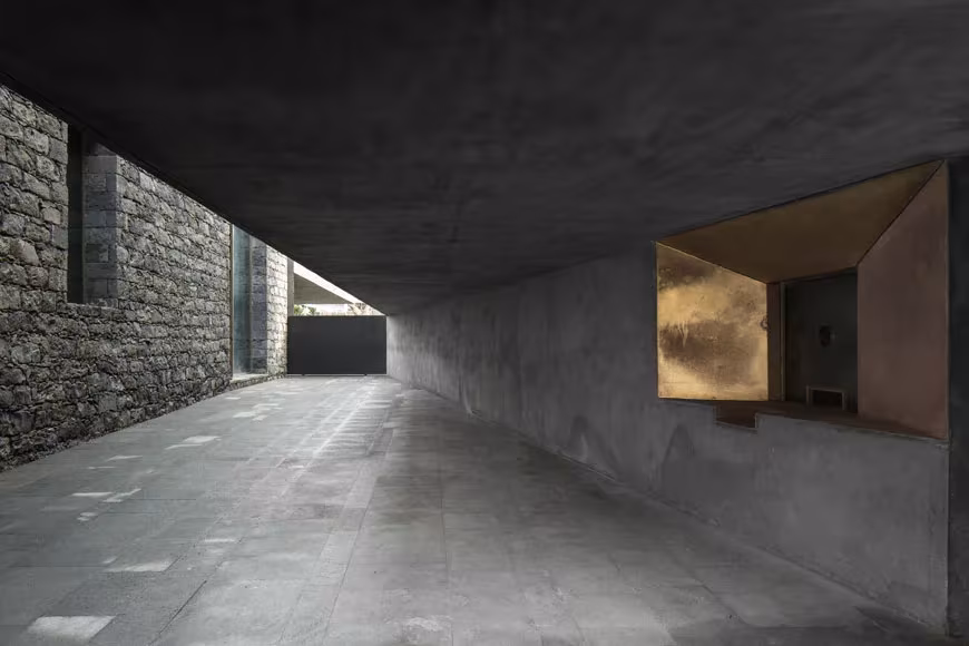 Pasaje cubierto de hormigón del centro Arquipélago junto a un muro de piedra, con una ventana revestida en dorado