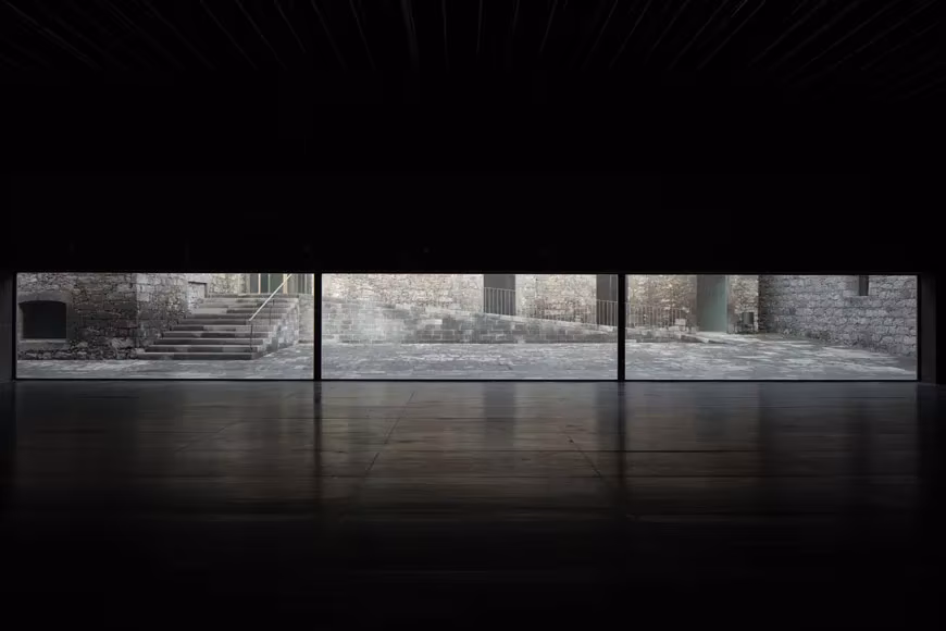 Sala oscura del centro Arquipélago con un ventanal horizontal que enmarca muros de piedra y escaleras exteriores