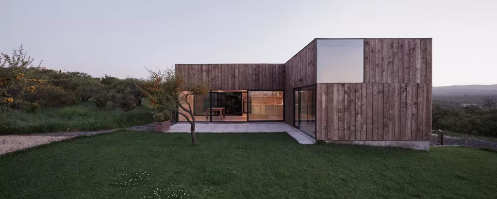 Casa CML en forma de L alrededor de un patio de césped con un árbol, con las ventanas iluminadas