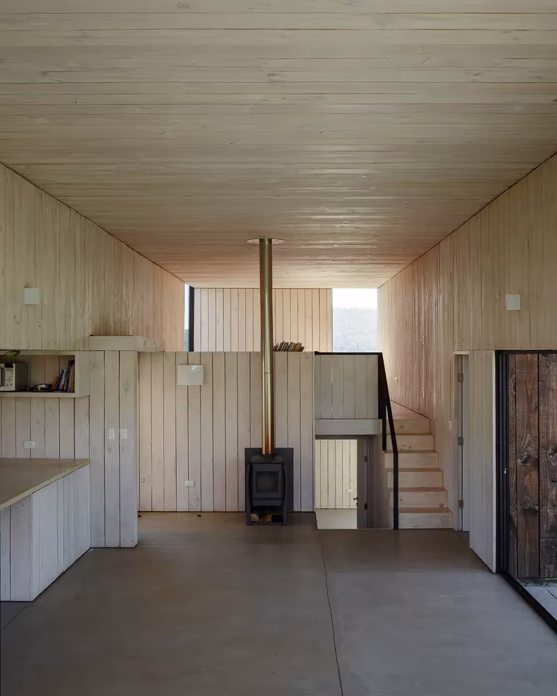 Interior de madera blanqueada de la Casa CML con estufa de leña, cocina y escalera al fondo