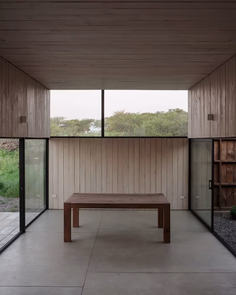 Comedor de la Casa CML con mesa de madera, suelo de hormigón y cerramientos de vidrio al campo
