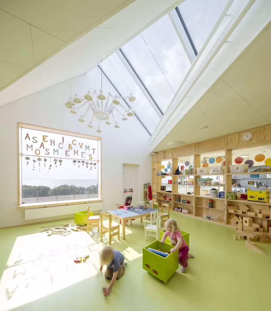 Dorte Mandrup. Guardería infantil Råå. Imagen interior Aula de la Guardería Råå con lucernario inclinado, suelo verde y niños jugando entre estanterías de madera