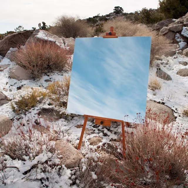 Espejo de Daniel Kukla en un caballete entre arbustos nevados que refleja un cielo azul con nubes