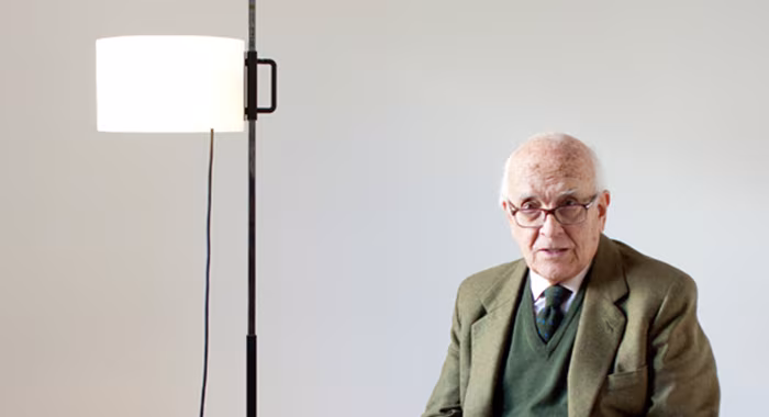 Retrato del diseñador Miguel Milá junto a su lámpara de pie TMM de pantalla blanca