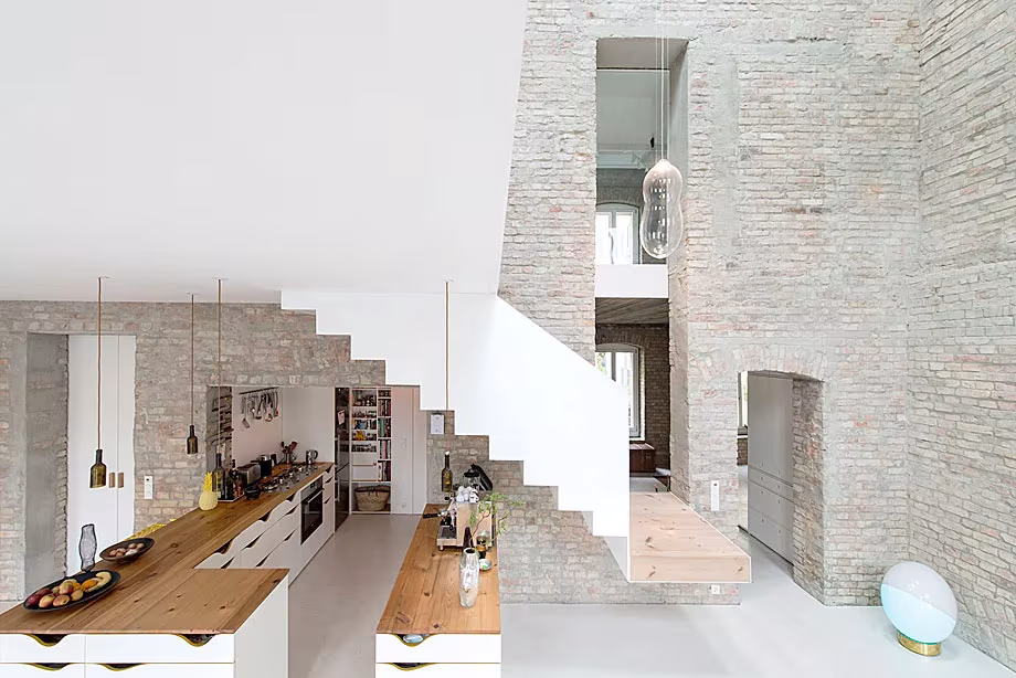 Cocina con encimera de madera bajo la escalera blanca y el muro de ladrillo visto de la Casa MMB