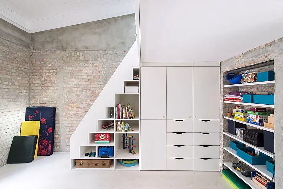 Escalera blanca con cajones, estantes y armarios integrados bajo los peldaños junto a un muro de ladrillo, en la Casa MMB