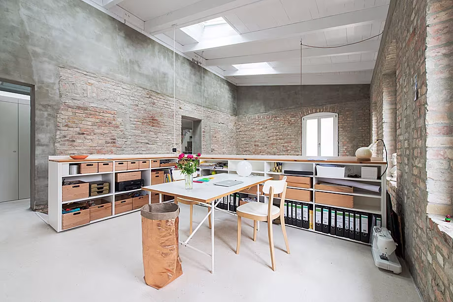 Estudio de trabajo con mesa blanca, estanterías bajas y muros de ladrillo bajo el lucernario, en la Casa MMB