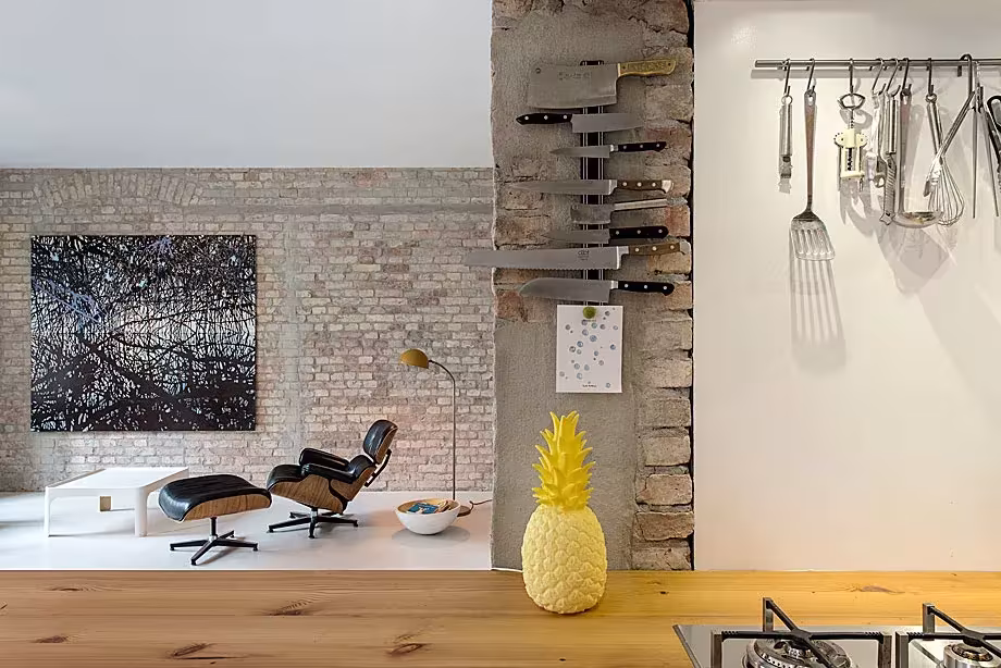 Cocina con encimera de madera, utensilios colgados y sillón de diseño ante un muro de ladrillo visto, en la Casa MMB