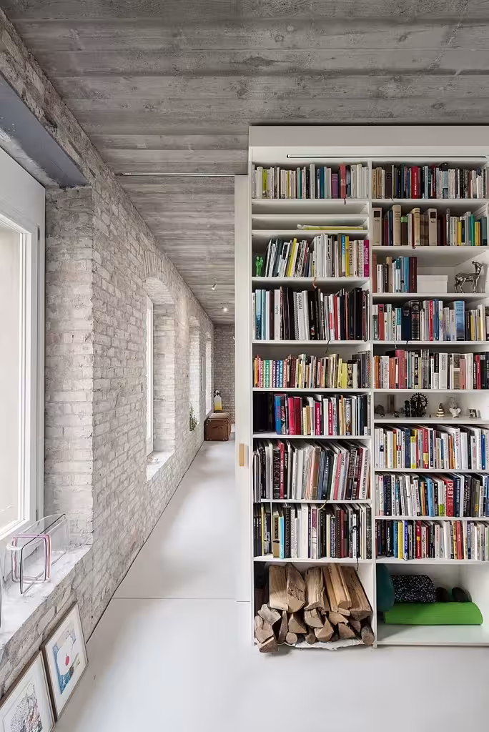 Pasillo con librería blanca, leña apilada y techo de hormigón visto junto a un muro de ladrillo en la Casa MMB