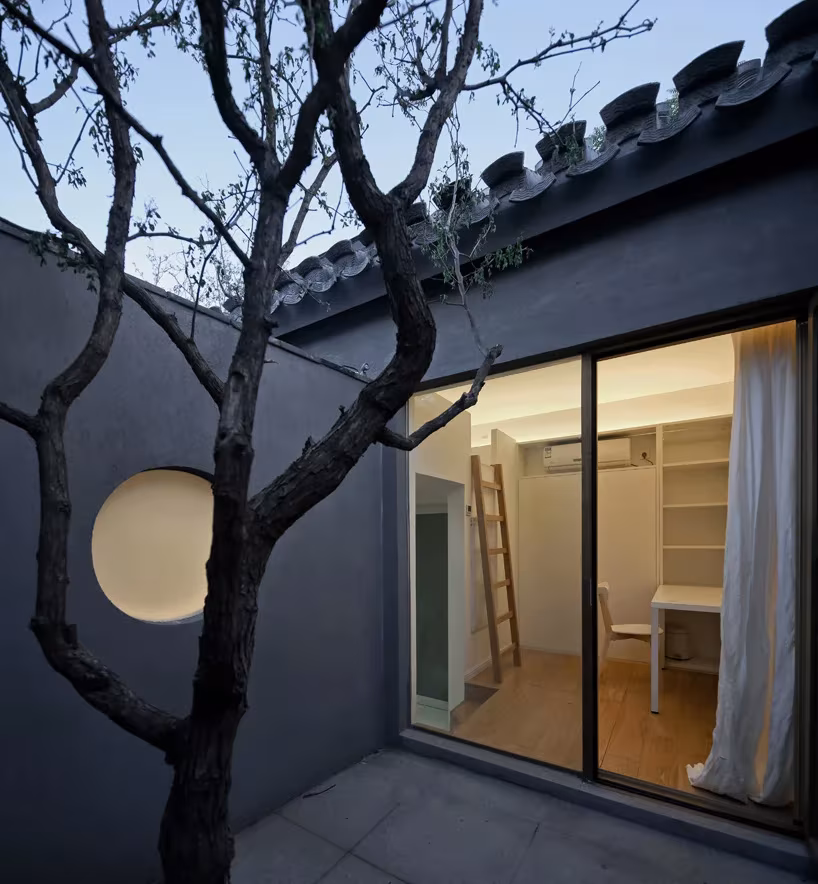 Split Courtyard House. TAO. habitación con patio privado Patio gris de la Split Courtyard House con un árbol, ventana circular y estudio iluminado tras el vidrio