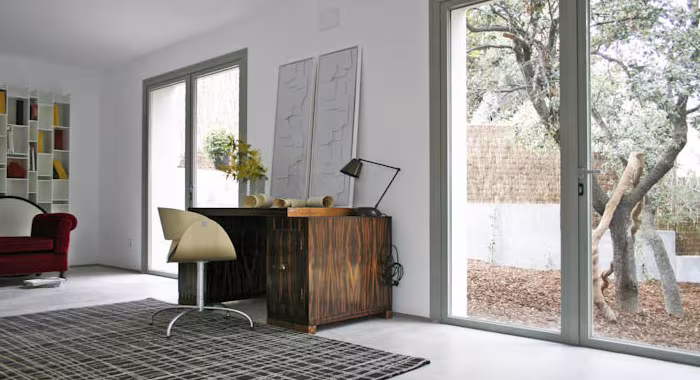 Escritorio de madera de estilo danés con silla tapizada junto a un gran ventanal abierto a un olivo