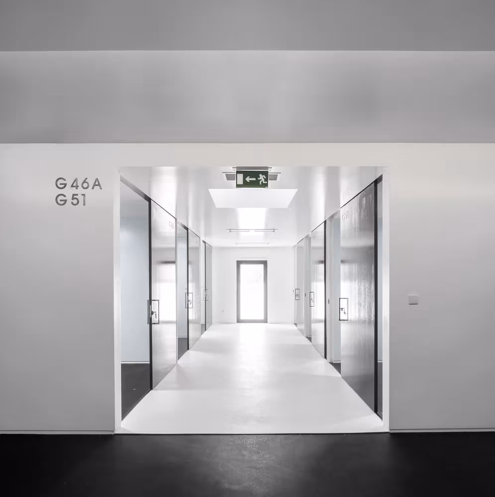 Pasillo de aulas de la ESTIG de Beja visto a través de un hueco, con puertas acristaladas y numeración en el muro