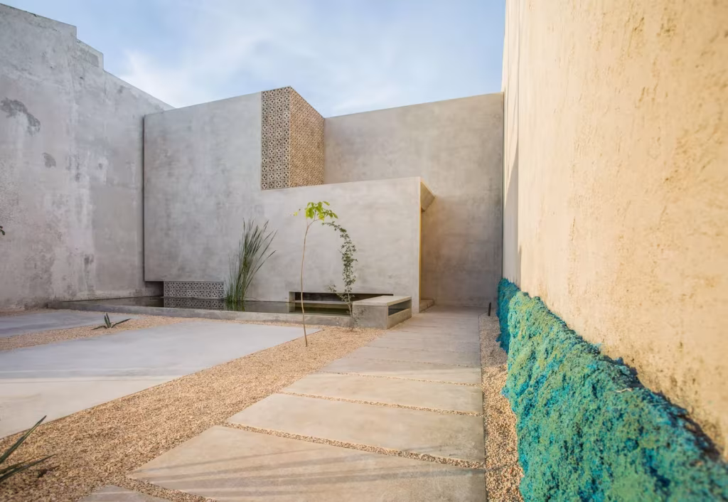 Patio de grava con losas, árbol joven y muro revestido de azul turquesa en la Casa Gabriela