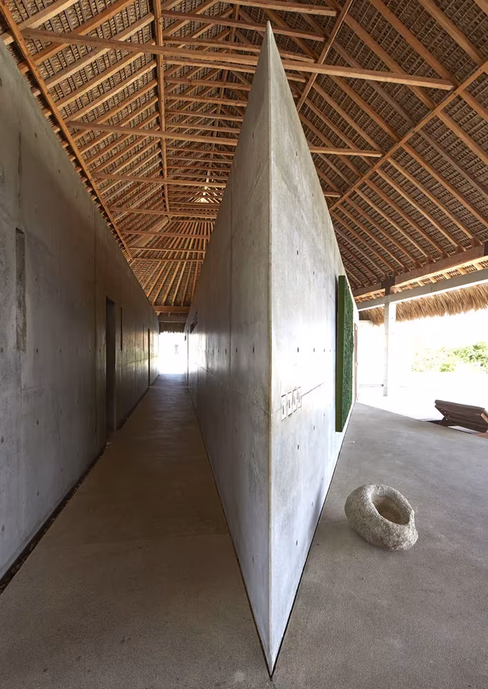 Galería interior de la Casa Wabi con un muro de hormigón inclinado y cubierta de palma sobre estructura de madera