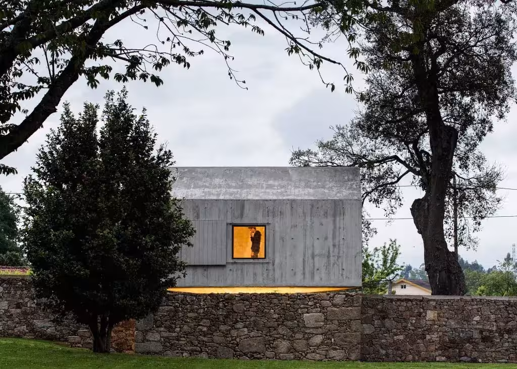The Dovecote por AZO Sequeira Arquitectos Associados. Casa de hormigón de The Dovecote asomando sobre el muro de mampostería, con una ventana iluminada y robles alrededor