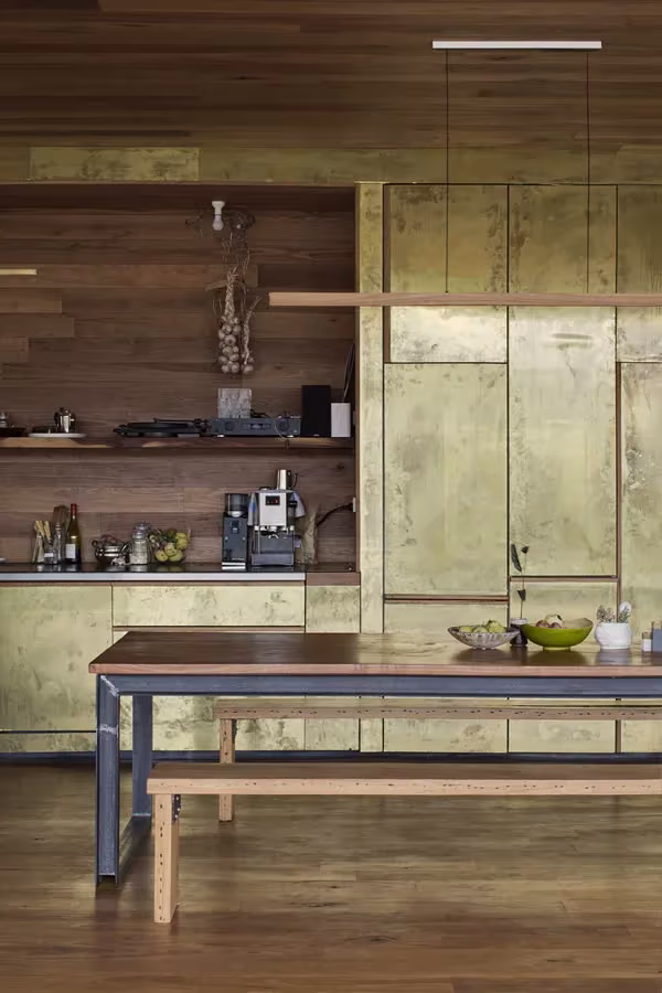 Cocina de la Sawmill House con paneles de latón envejecido, estantes de madera y mesa con banco