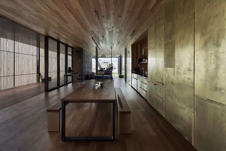Sala alargada con mesa de madera y acero, muro dorado de latón y techo de madera en la Sawmill House