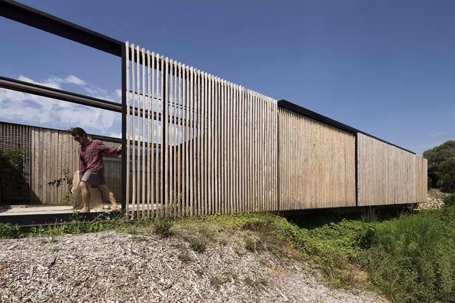 Fachada de lamas de madera de la Sawmill House, con una persona abriendo un panel junto a la gravilla