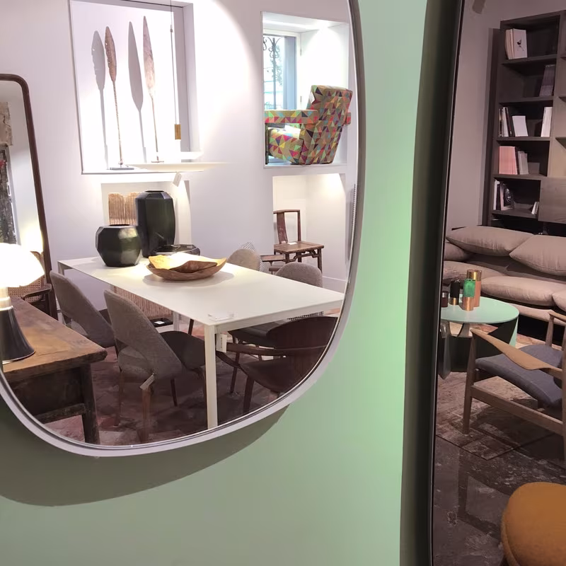 Espejo ovalado en una pared verde que refleja un comedor con mesa blanca y sillas de madera