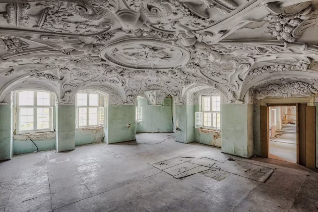 Salón abandonado con techo de estuco barroco, paredes verdes desconchadas y ventanas en arco